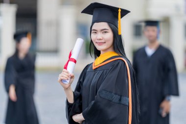 Mezuniyet elbisesi ve havan tahtasıyla güzel bir Asyalı kadın üniversite mezunu üniversite mezuniyetine katıldıktan sonra üniversite binasının önünde diploma tutuyor.