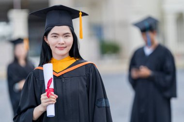 Mezuniyet elbisesi ve havan tahtasıyla güzel bir Asyalı kadın üniversite mezunu üniversite mezuniyetine katıldıktan sonra üniversite binasının önünde diploma tutuyor.