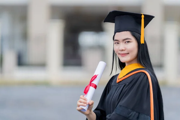 卒業生のガウンとモーターボードを着た若い美しいアジアの女性大学卒業生は 大学の卒業式に参加した後 大学の建物の前に学位証明書スタンドを保持しています — ストック写真