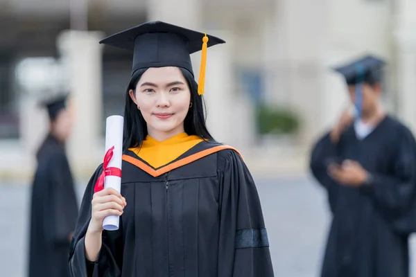 卒業生のガウンとモーターボードを着た若い美しいアジアの女性大学卒業生は 大学の卒業式に参加した後 大学の建物の前に学位証明書スタンドを保持しています — ストック写真