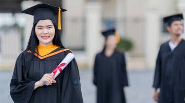 Mezuniyet elbisesi ve havan tahtasıyla güzel bir Asyalı kadın üniversite mezunu üniversite mezuniyetine katıldıktan sonra üniversite binasının önünde diploma tutuyor.