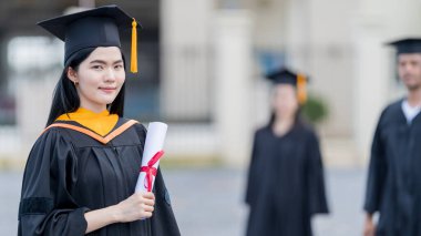 Mezuniyet elbisesi ve havan tahtasıyla güzel bir Asyalı kadın üniversite mezunu üniversite mezuniyetine katıldıktan sonra üniversite binasının önünde diploma tutuyor.