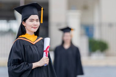 Mezuniyet elbisesi ve havan tahtasıyla güzel bir Asyalı kadın üniversite mezunu üniversite mezuniyetine katıldıktan sonra üniversite binasının önünde diploma tutuyor.