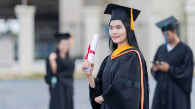 Mezuniyet elbisesi ve havan tahtasıyla güzel bir Asyalı kadın üniversite mezunu üniversite mezuniyetine katıldıktan sonra üniversite binasının önünde diploma tutuyor.