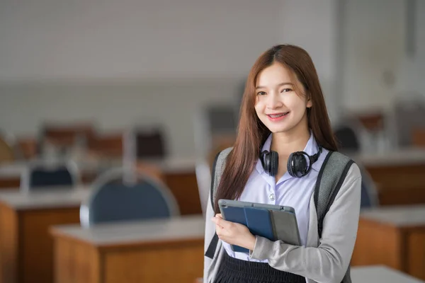 Portrait of young Asian woman student in uniform holding tablet in smart and happy pose in university or college classroom. Youth girl student and tutoring education with technology learning concept.