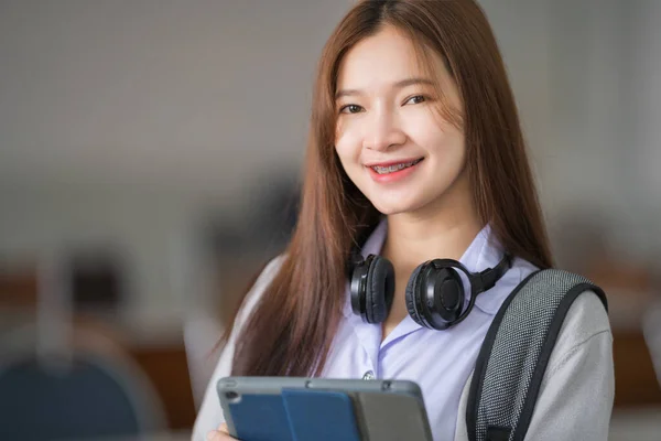 Portrait of young Asian woman student in uniform holding tablet in smart and happy pose in university or college classroom. Youth girl student and tutoring education with technology learning concept.
