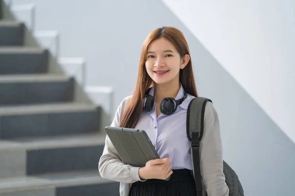Portrait of young Asian woman student in uniform holding tablet in smart and happy pose in university or college classroom. Youth girl student and tutoring education with technology learning concept