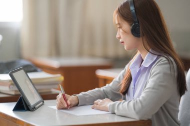 Kablosuz kulaklıklı genç Asyalı bir üniversite öğrencisi tabletteki hocayı dinliyor ve not defterine tek başına ders çalışması için yazıyor.