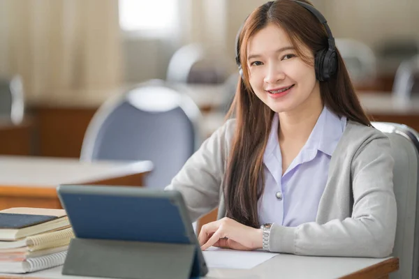 Young Asian woman college student with wireless headphones watches and listens to the lecturer on tablet and writes on the notebook to study online by herself in classroom
