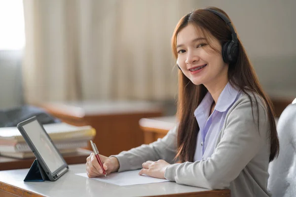 若いアジアの女性の大学生は ワイヤレスヘッドフォンの時計とタブレット上の講師に耳を傾け 教室で自分でオンラインで勉強するためにノートに書き込みます — ストック写真
