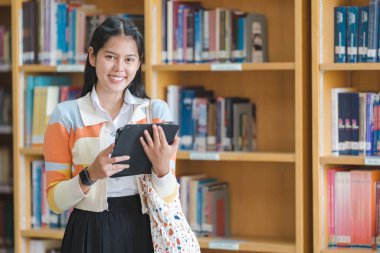 Üniversite ya da üniversite kütüphanesinde elinde akıllı ve mutlu pozlar veren üniformalı genç bir Asyalı kız öğrenci. Genç kız öğrenci ve teknolojik öğrenim konsepti ile özel eğitim