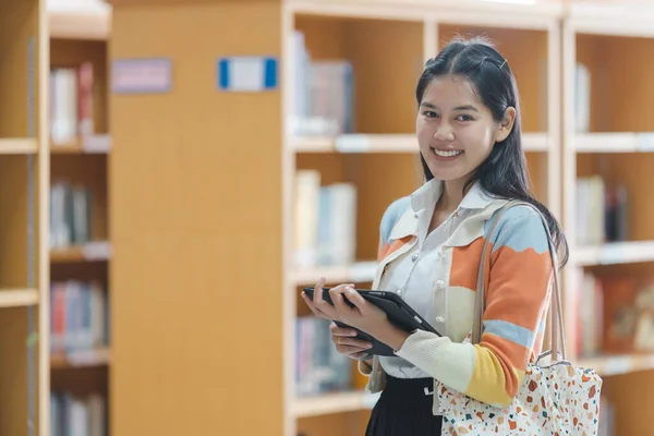 Young Asian woman student in uniform holding a tablet in smart and happy pose in university or college library. Youth girl student and tutoring education with technological learning concept