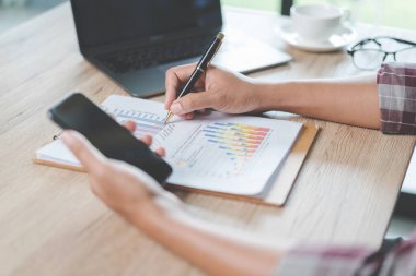 Yakın plan iş adamı elinde kalem, akıllı telefon ve finansal grafiği işaret ederek ahşap masadaki iş raporlarını evde bilgisayarlı dizüstü bilgisayarla kontrol ediyor. Stok fotoğrafı