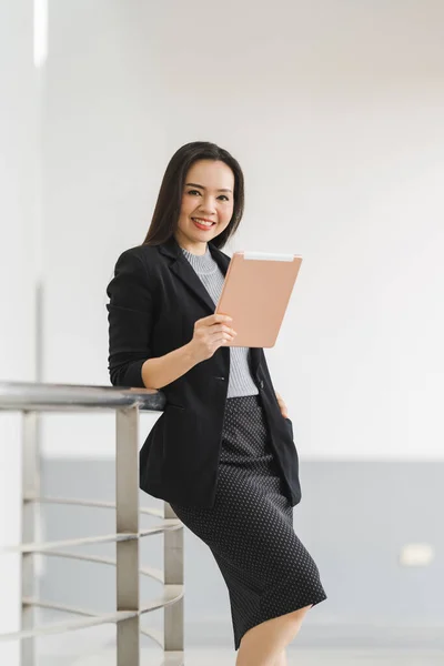 ビジネススーツ姿の明るいアジアの実業家が ビジネスビルのデジタルタブレットを使って立っている姿を描いています 業務用ストックフォト — ストック写真
