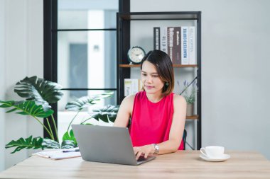 Evde rahat elbiseler giymiş neşeli, orta yaşlı Asyalı bir iş kadını, bilgisayar laptopunda e-postaları kontrol ediyor, finansal muhasebe belgelerine yazı yazıyor. İş yeri fotoğrafı