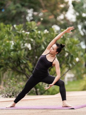 Spor kıyafetli orta yaşlı, kendinden emin bir Asyalı kadın sabahları arka bahçede yoga minderinde yoga egzersizi yapıyor. Genç bir kadın doğa parkında yoga egzersizi yapıyor.