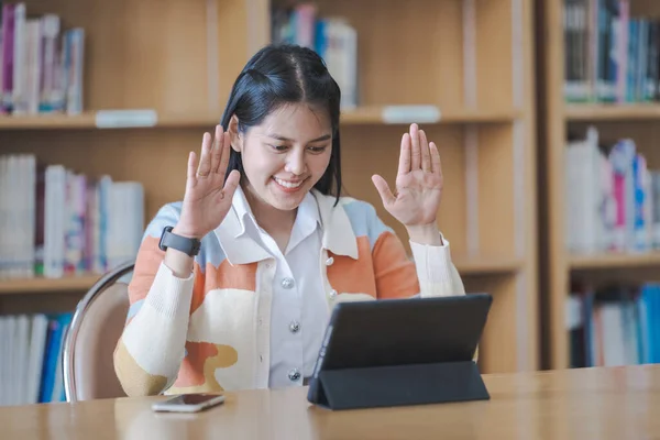 Young woman Asian college student in student uniform studying online, reading a book, using digital tablet or laptop in university library while classroom being restricted during COVID-19 pandemic