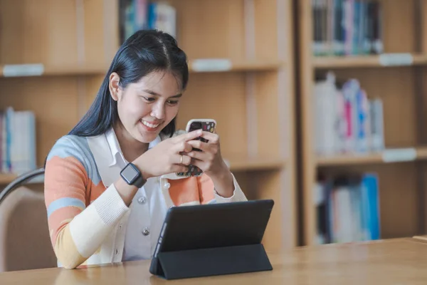 Young woman Asian college student in student uniform studying online, reading a book, using digital tablet or laptop in university library while classroom being restricted during COVID-19 pandemic