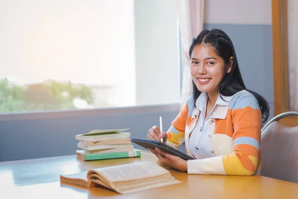 A young happy woman Asian college student in student uniform studying online, reading a book, using digital tablet or laptop in the university library in free time by oneself