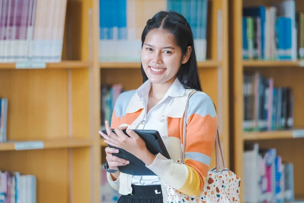 A young happy woman Asian college student in student uniform studying online, reading a book, using digital tablet or laptop in the university library in free time by oneself