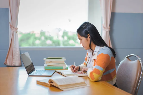 A stressful Asian college woman student in student uniform studying online, reading a book, using digital tablet or laptop in university library in free time by oneself