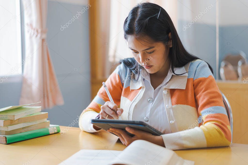 A stressful Asian college woman student in student uniform studying online, reading a book, using digital tablet or laptop in university library in free time by oneself