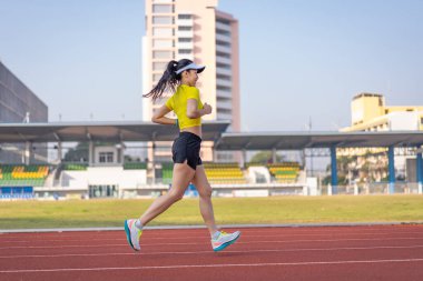 Genç bir Asyalı bayan atlet, formda ve sağlıklı yaşam tarzını korumak için güneşli bir sabahta şehir stadyumunda koşu pistinde koşuyor. Genç fitness kadını stadyum pistinde koşuyor. Spor ve eğlence.
