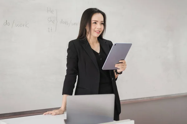 教室で現代語を教えるために デジタルタブレットとラップトップで統一された黒のビジネススーツで自信を持って陽気なアジアの女性教師のストックフォト肖像画 — ストック写真