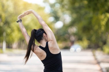 Spor kıyafetleri içinde neşeli bir Asyalı kadın koşucu, akşam üstü gün batımında şehir parkında koşuya çıkmadan önce esneme hareketleri yapıyor.