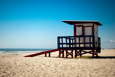 Canlı bir cankurtaran kulesi, güneşli bir sahilde gururla durur yumuşak kum, nazik dalgalar ve berrak mavi bir gökyüzü tarafından çerçevelenmiştir..