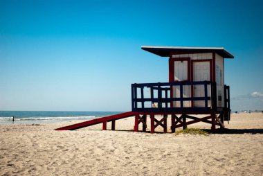 Canlı bir cankurtaran kulesi, güneşli bir sahilde gururla durur yumuşak kum, nazik dalgalar ve berrak mavi bir gökyüzü tarafından çerçevelenmiştir..