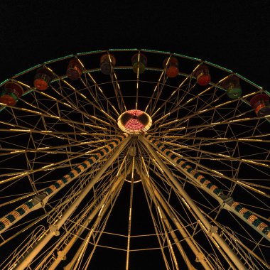 Yüksek bir dönme dolaba bakan dramatik alçak açılı çekim karanlık bir gökyüzüne doğru, parlak bir şekilde aydınlatılmış sıcak altın ışıklarla. Bir karnaval ya da panayır alanının ikonik süslemesini yakalamak.