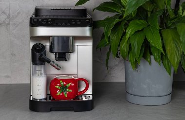Çağdaş kahve makinesi, yakındaki yemyeşil bir bitki tarafından tamamlanmış parlak bir tezgahın üzerinde canlı kırmızı bir kupa ve süt kabını barındırıyor.