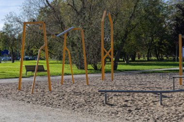 Modern oyun alanı ekipmanları, verimli çimenler ve kumlu zemini olan güneşli bir parkta yer alan salıncaklar ve jimnastik halkaları içerir.