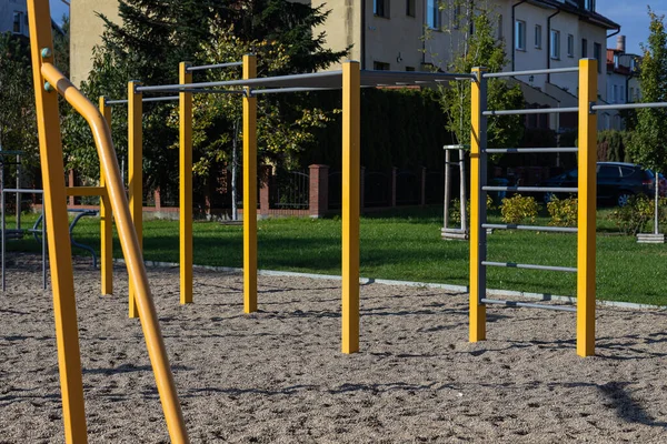 Oyun parkı ekipmanları, çimenli alan ve ağaçlarla çevrili sarı maymun parmaklıklarını içeriyor ve çocuklar için açık hava eğlencesi alanı yaratıyor.