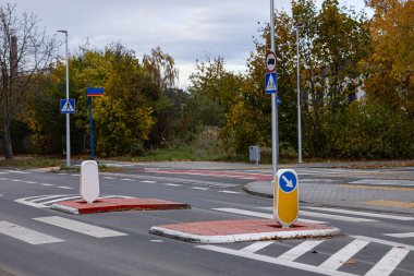 Renkli sonbahar yeşillikleriyle çevrili çeşitli trafik işaretleri ve yaya geçişleriyle şehir yolu kesişmesi, kentsel altyapının ve güvenlik tedbirlerinin önemini gösteriyor