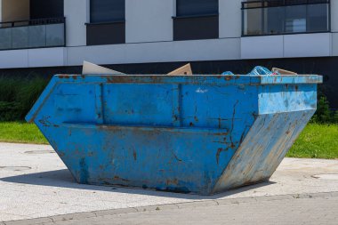 Güneşli bir günde modern konut binasının yakınına yerleştirilmiş büyük mavi metal inşaat çöplüğü, yenileme enkazı ve kentsel temizlik konteynırı