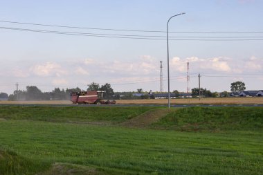 Kırsal bir yolun yakınındaki gün batımında altın buğday tarlasında çalışan kırmızı hasatçıyı birleştirir. Tarım makineleri olgun tahıl ürünleri topluyor. Endüstriyel tarım ve gıda üretimi
