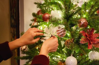 Bir kadının elleri yeşil yapay Noel ağacına bir süs takar, kırmızı, altın süsler ve yeşil dallar arasında parlayan küçük beyaz ışıklarla süslenir. Odak noktası süs.