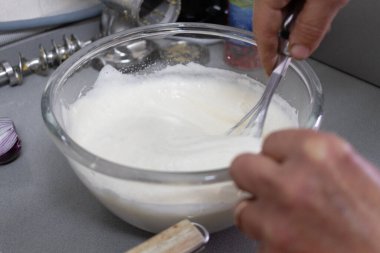 Yaşlı ellerin, gri mutfak tezgahında büyük bir cam kasede kalın beyaz krema çırpması, metal çatal bıçaklar, mutfak mikser parçaları ve gün ışığında yakınlarda duran dilimlenmiş kırmızı soğan..