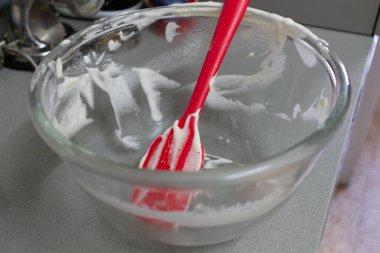 Beyaz hamurlu cam bir kase ve içinde kırmızı silikon bir spatula dinleniyor, gri mutfak tezgahında oturuyor ve mutfak arka planında bulanık pişirme aletleri var..