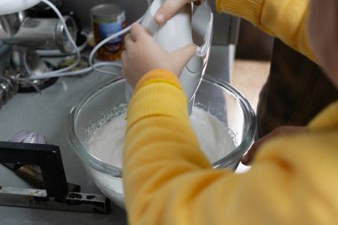 Sarı kazaklı küçük bir çocuk yemek yapmayı öğreniyor, bir yetişkin ellerini elektrikli bir mikserle karıştırıyor, mutfak tezgahında cam bir kasede kremalı hamuru kırbaçlıyor, evde takım çalışması yapıyor..