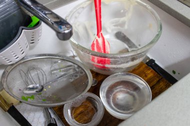 Musluğun altındaki kirli çatal bıçaklarla dolu kirli mutfak lavabosu: cam karıştırma kabı kırmızı spatulayla, metal elek kaşıkla, çırpma bıçağıyla, süngerle ve ıslaklık tahtasındaki yemek artıklarıyla..