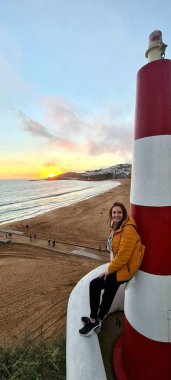 Gülümseyen bir kadın, kırmızı beyaz çizgili bir deniz fenerinin yanında oturmuş sahildeki büyüleyici günbatımını seyrediyor. Dalgalar yavaşça altın kuma çarpar..