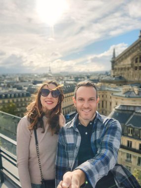Güneşli bir günde arka planda Eyfel Kulesi olan ve panoramik manzaralı bir balkonda selfie çeken gülümseyen bir çift..