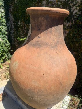 Köy yüzeyi ve aşınmış dokusu ile antik bir kil vazo, yeşil bitki örtüsü ve güneş ışığıyla çevrili doğal bir ortamda, gelenek ve tarih duygusunu aktarıyor..