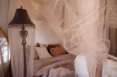 A cozy bedroom with pillows and a beige fluffy bedspread. A canopy headboard. The bedroom interior features a bed and a sitting area. A vintage lamp and a round mirror
