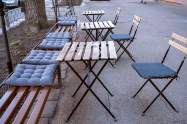 An empty outdoor seating area in early spring or late fall. The outdoor seating composition features chairs and benches with cushions and wooden tables. A wooden bench sitting in a chair.