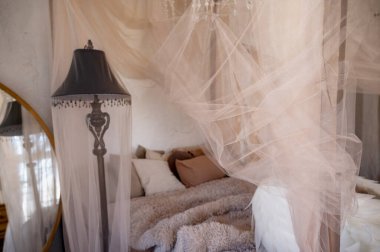 A cozy bedroom with pillows and a beige fluffy bedspread. A canopy headboard. The bedroom interior features a bed and a sitting area. A vintage lamp and a round mirror