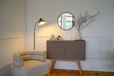 Sıcak ışık saçan bir masa lambasıyla dekoratif bir köşe manzarası. Beyaz duvarların karşısında açık renkli bir koltuk, kahverengi bir çekmece, duvarda bir ayna, bir vazo ve kitaplar var..